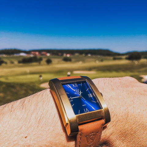 Herrklocka i borstat guld med blå romersk urtavla - närbild på fyrkantig klocka med golfbana i bakgrunden
