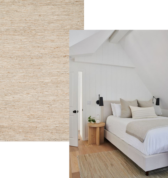 a white bedroom with a sloped wooden roof. A natural jute rug lies beneath a bed made up with pale linen bedding. A light wooden bedside table has a small plant on it and a small black pendant light sits just above it 