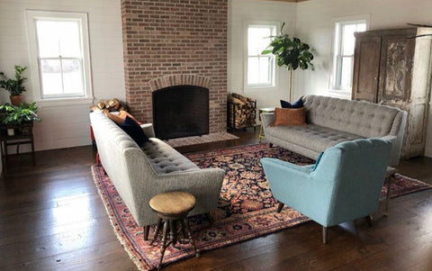 a predominantly brown living room, with an exposed brick fireplace, dark floorboards and large wooden cupboard. There are two light brown sofas and a blue/grey arm chair sat upon an antique rug. A wood pile and potted plant are visible in the far right hand corner