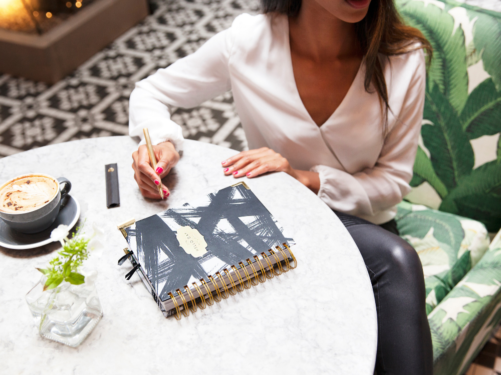 Woman sitting at table with coffee and a diary, writing. 