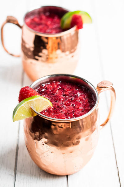 two copper mugs filled with pink liquid and a raspberry and sliced lime on its rim