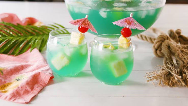 two clear glass filled with bluish green liquid with pineapple skewers and tiny umbrellas