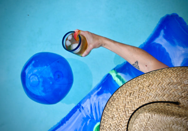A person holds a glass of cold brew iced tea while lounging on a raft in a pool. 