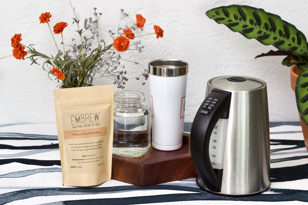 An electric tea kettle, a pouch of Embrew tea, some flowers, a glass of water, and a mug sit on a counter. 