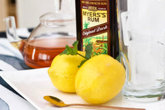 Two lemons sit on a plate beside a teaspoon, a glass of rum, and a tea kettle. 
