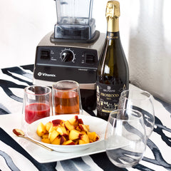 A dish full of apple pieces sits on a counter next to two beverage glasses, a bottle of wine, and a blender. 