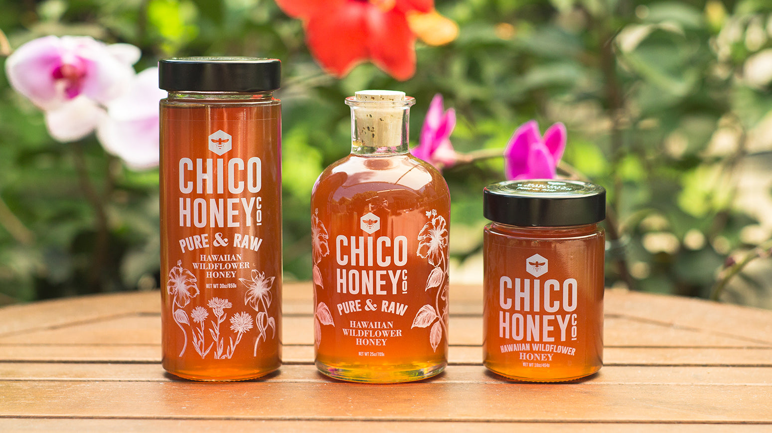 Three different sizes of OHB Hawaiian Wildflower Honey from large to small on a light wood table with Hawaiian flowers in background