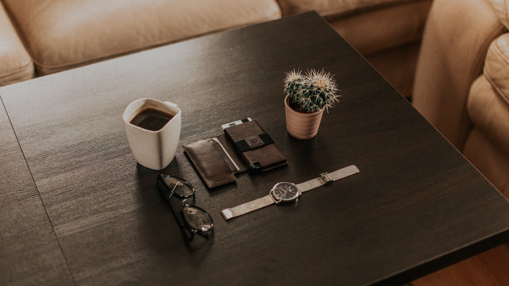 Background is a dark wood table with a few items neatly arranged in the center. The items are an Ekster trackable smart wallet, a watch, a plant, a mug of coffee, and a pair of sunglasses.