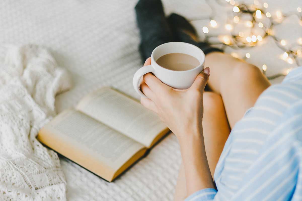 woman in bed reading a book with cup of coffee