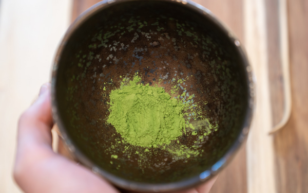 high resolution matcha green tea in a brown matcha bowl, a white skinned hand holds it above a wooden deck