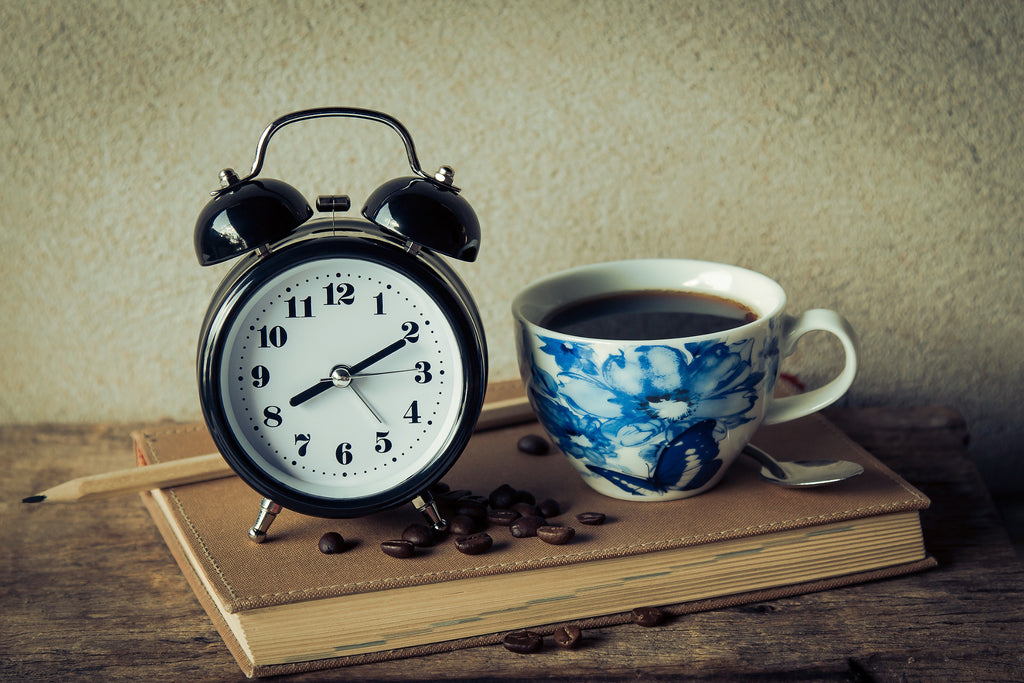 analog alarm clock next to coffee in mug with blue flower sitting on top of book 