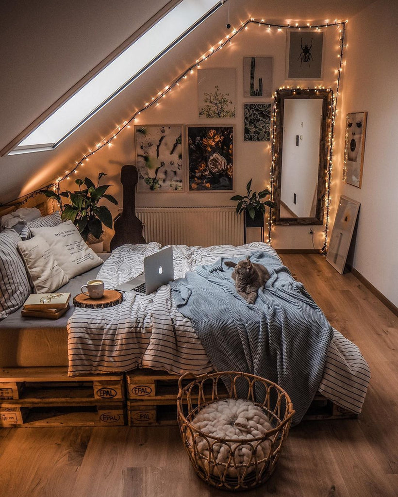 Overview of bedroom with fairy lights, bed with striped sheets, books, and a grey cat.