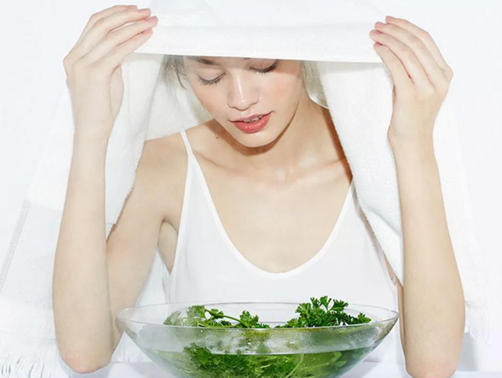 Woman holding towel above her as she steams her face above glass bowl with green vegetables.