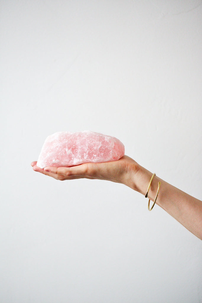 Hand holding a palm-sized rose quartz stone in front of white wall background.