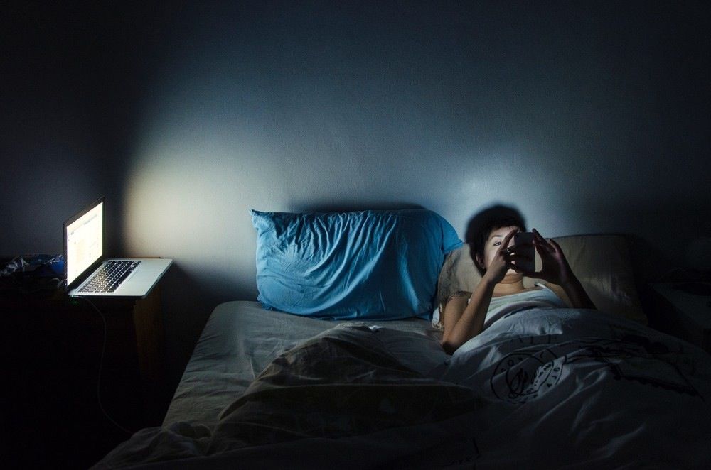 Woman in the dark, in bed, on her phone with a laptop open beside them.