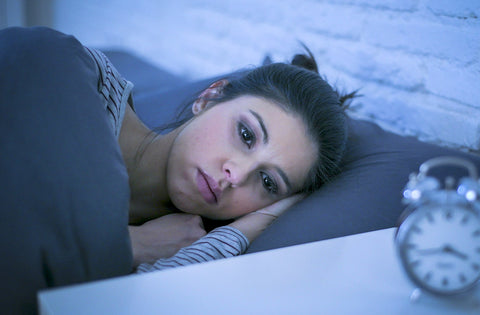 photo of a woman laying in bed with a sad look on her face. An alarm clock is in the foreground.