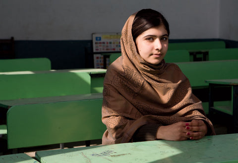 Malala Yousafzai with headscarf on sitting at a green desk with hands clasped.