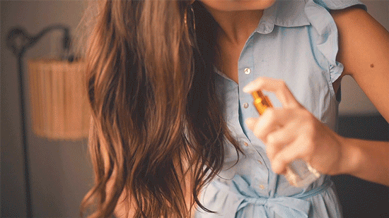 A woman with long brown hair in a blue shirt dress spraying DefineMe Hair Fragrance Mist into her hair.