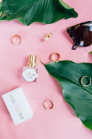 DefineMe Fragrance Clara Mist lying on pink background with fragrance box, gold rings, and sunglasses.