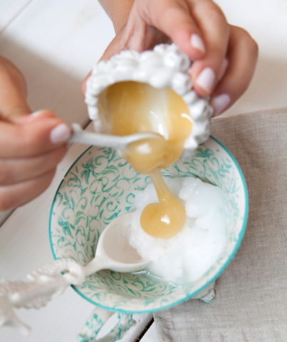 Pouring honey with a spoon into green and white bowl with coconut oil.