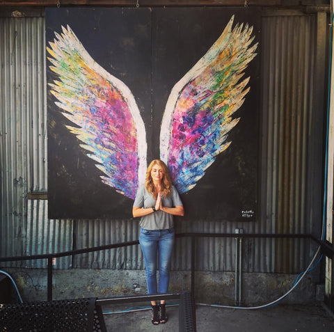 DefineMe Fragrance Founder, Jennifer McKay Newton standing in front of colorful painted wings on a wall and holding hands in prayer.