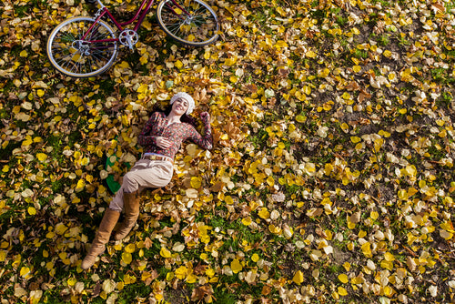 Woman lying in field of yellow leaves with bicycle on side.