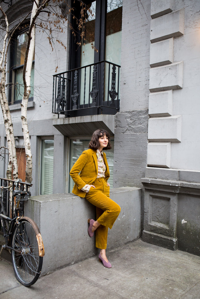 Image of Alyssa Coscarelli in a mustard corduroy ensemble leaning against a concrete wall. 