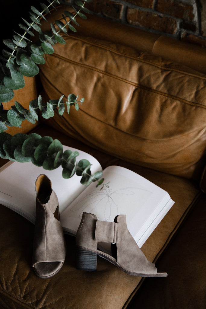 The Coclico Zephira Sandal on a leather couch with eucalyptus plant and opened book in background. 