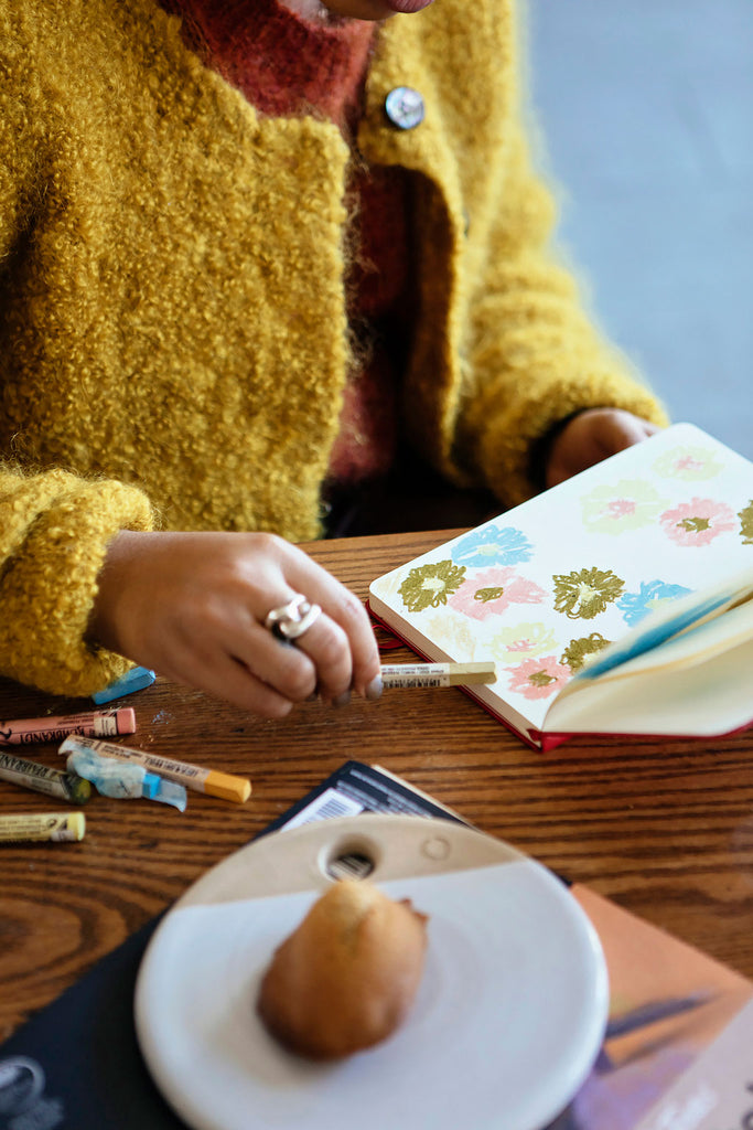 Close up of Elise Peterson drawing with her art materials scattered across the wooden table.