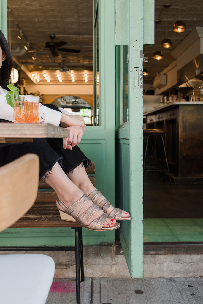 Close up of Giorgia Zedda sitting on a wooden bench showing off her O'Boy Sandal in Rose Metallic Leather.
