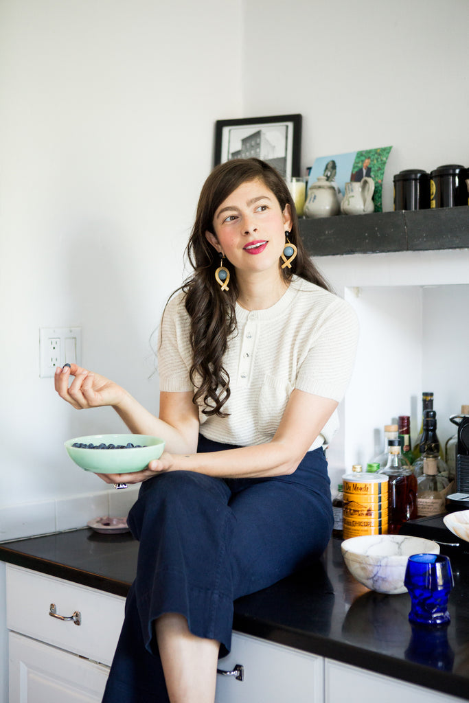 Sunlit picture of Erin Allweiss seated on the kitchen counter holding a bowl of blueberries. 