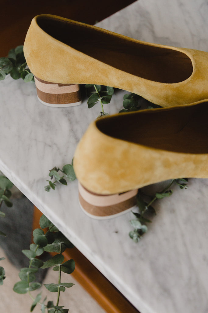 Close up of the Epic Heel in Butterfly Yellow suede placed on a marble table beside plants. 