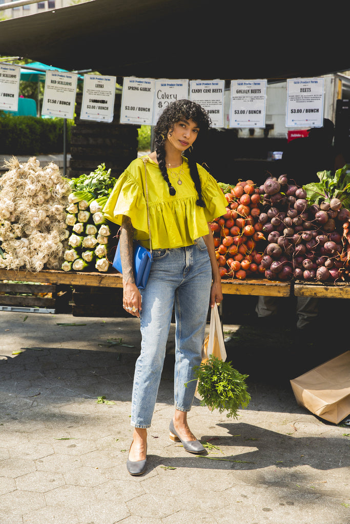 Full-body shot of Sophia Roe in front of a vegetable stand carrying a bag of groceries and wearing the Epic Heel. 