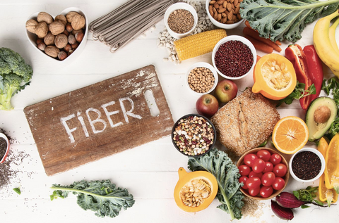 A flatlay image of wooden chopping board with a word fiber, surrounded by different food such as Tomato, nuts, peppers, banana, orange and avocado