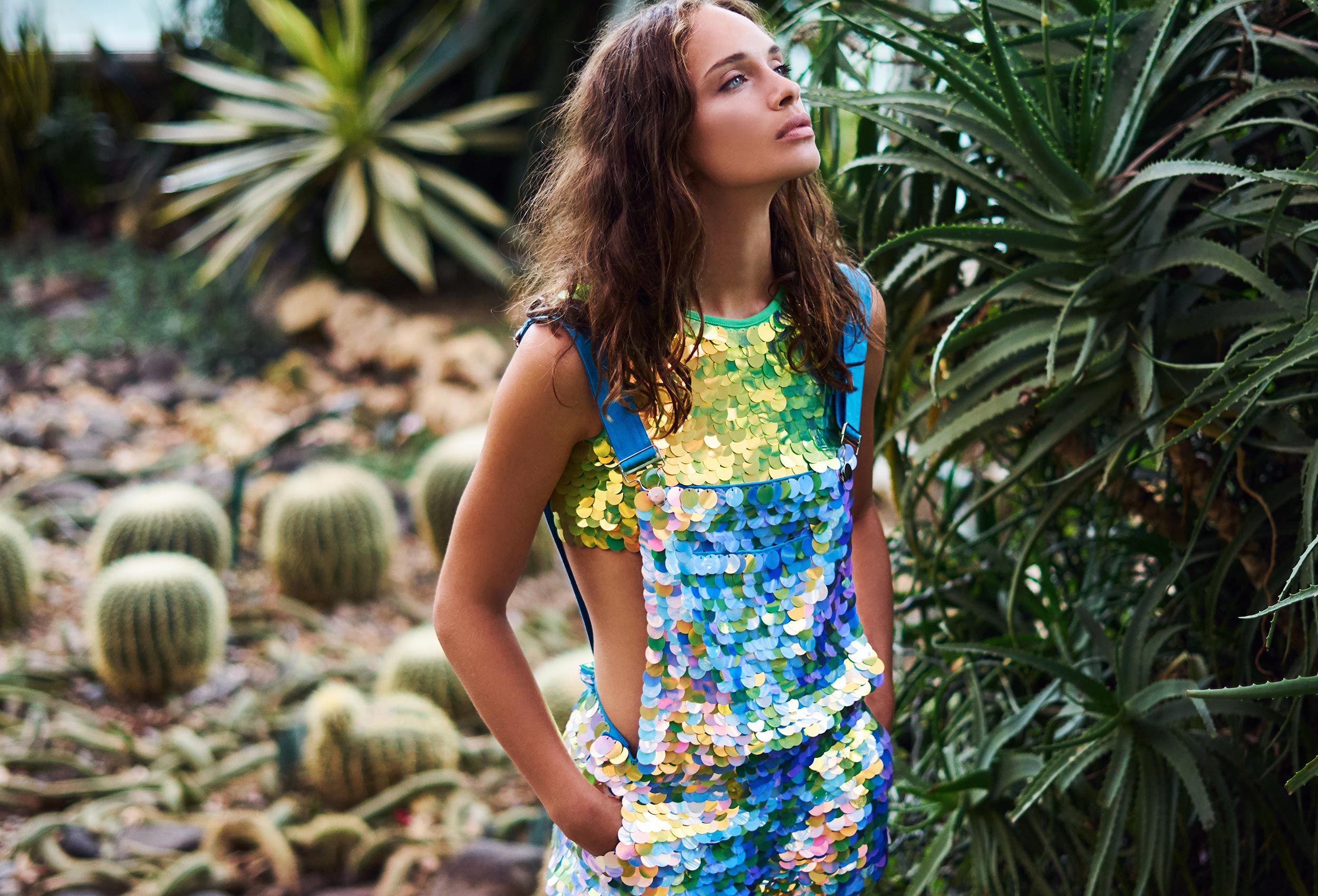 woman wearing dungaree shorts and a crop top covered in large round iridescent green and blue sequins standing in an old glasshouse surrounded by foliage