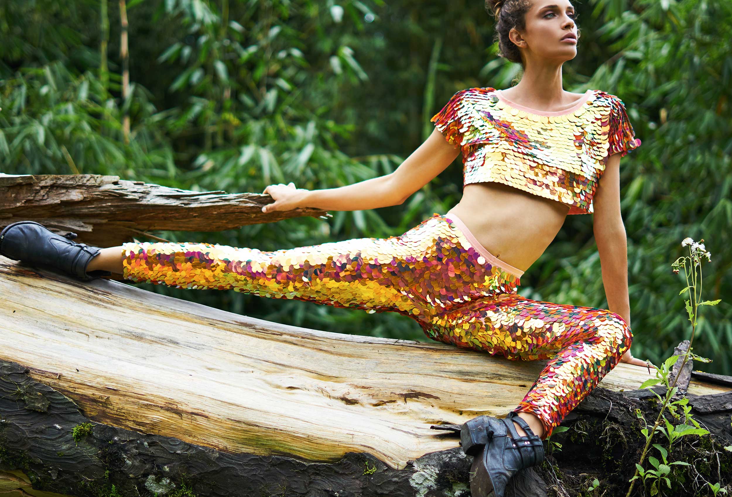 woman wearing iridescent red sequin leggings and cropped t-shirt doing the splits with one knee bent on a large fallen tree trunk