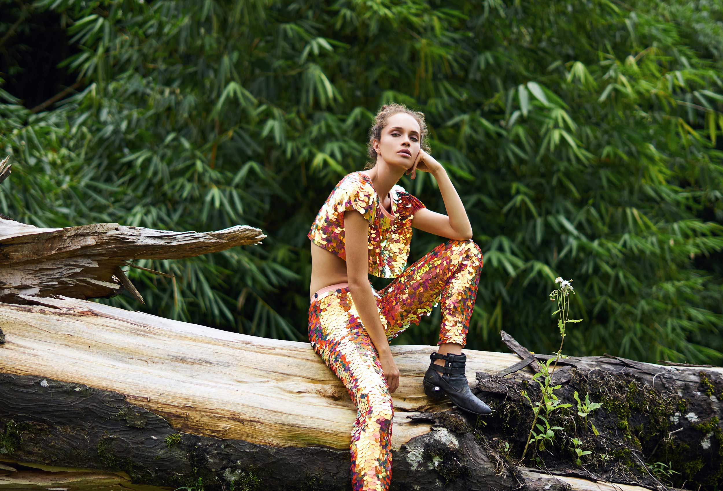 woman in leggings and a cropped t-shirt covered in large red iridescent sequins sitting on a large fallen tree