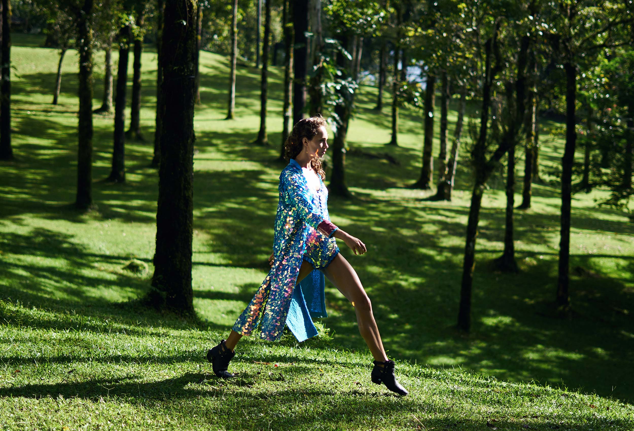 woman in a long kimono covered in large round blue iridescent sequins taking a big stride amongst trees in a grassy landscape