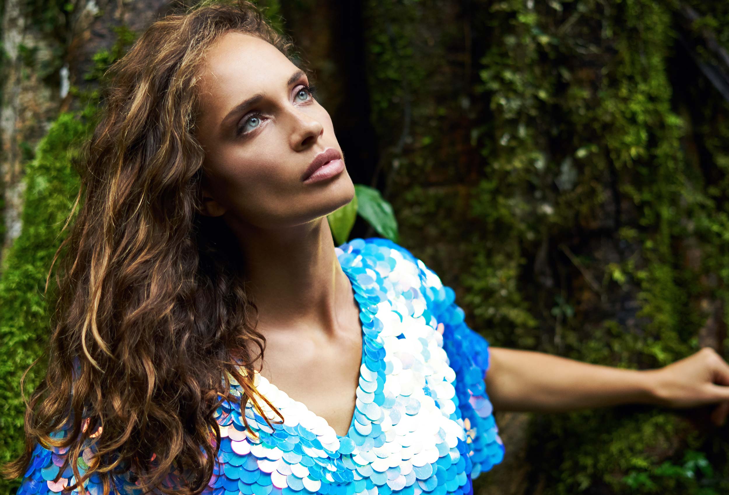 woman looking into the distance with dark foliage behind wearing a v neck top covered with large round blue sequins