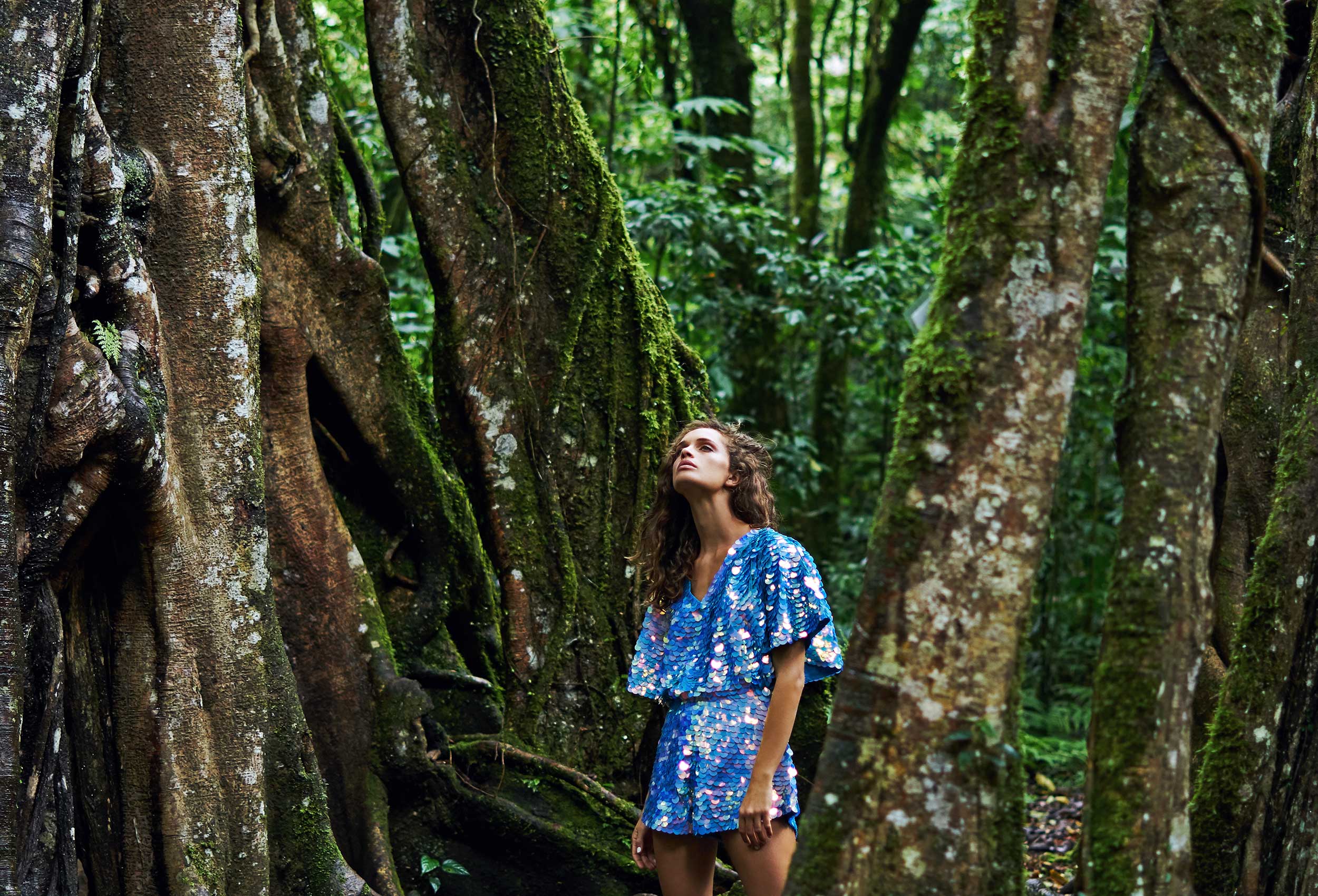 woman wearing a blue sequin covered romper playsuit with cape sleeves looking upwards standing amongst huge trees