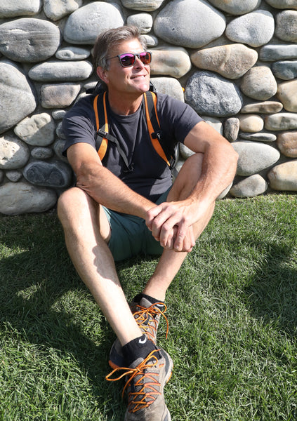 Older man in hiking gear sitting down and smiling while wearing a red multi-color mirrored sunglasses.