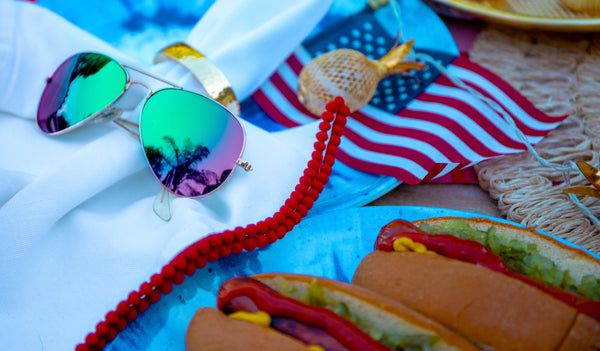 Green mirrored sunglasses next to grilled hot dogs and the American flag in the background.
