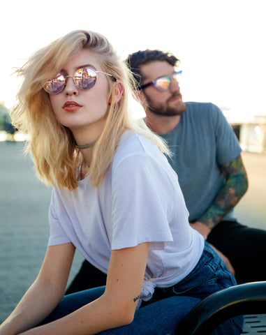 Woman and man wearing rose gold sunglasses outside, looking in opposite directions.