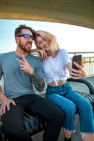 Man and women sitting on a bench wearing blue light lenses taking a selfie while making a funny face