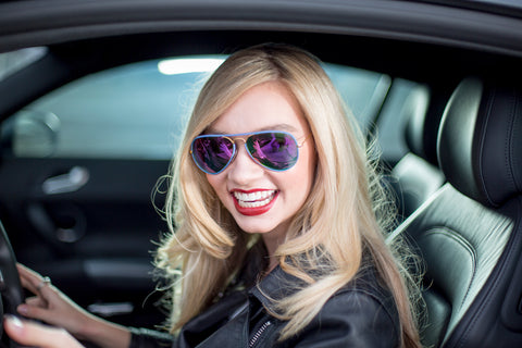 Blonde women in a car smiling while wearing a leather jacket and purple mirrored sunglasses.