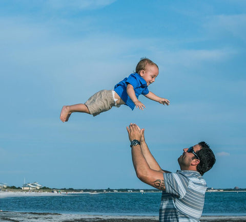 A dad playing with his toddler son, on the beach, by throwing him up and catching him. Photo taken by David Martin Photography.