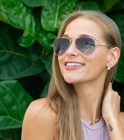 Smiling blonde woman wearing aviator sunglasses with grey lenses in front of greenery