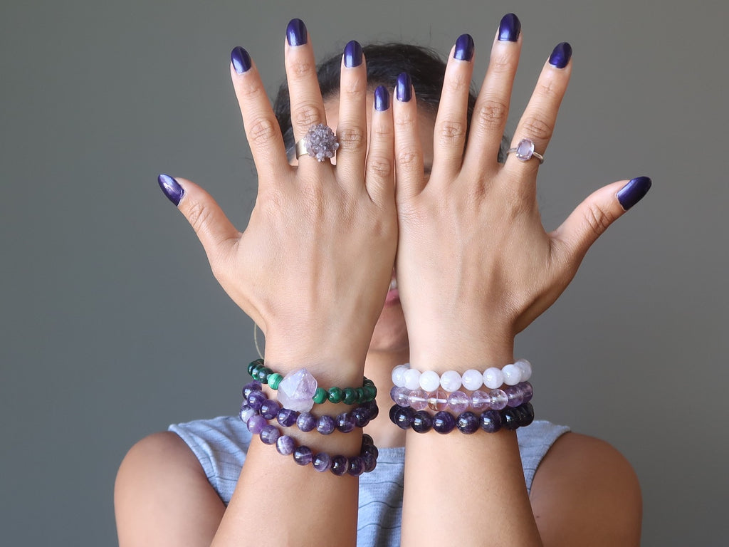 hands in front of face wearing different types of amethyst stretch bracelets and rings