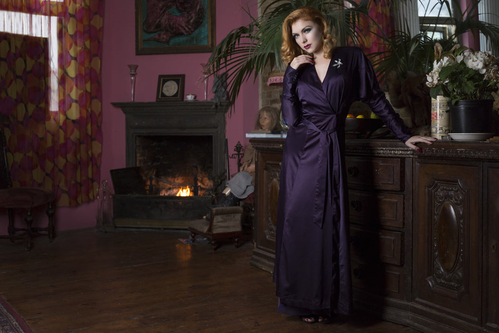 A model in a plum satin full length 1930's style lounges against a fireplace and wooden dresser