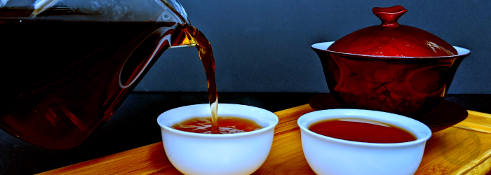 pu-erh tea is served from a glass pitcher into two white tasting cups in front of a red gaiwan